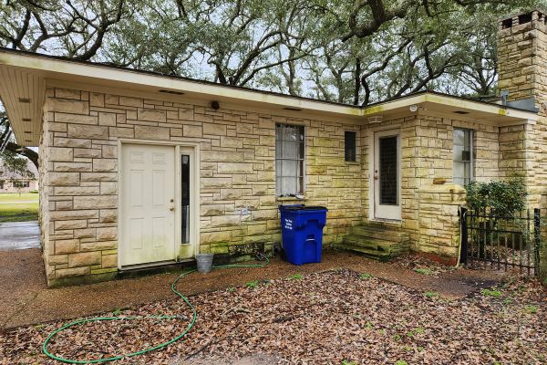 A section of a house's exterior with visible dirt, stains, and discoloration on the stone surface, and the ground covered with fallen leaves and dirt.