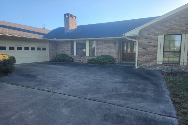 The driveway of a house showing a slightly dirty and uneven surface, with stains and patches of discoloration, as well as some overgrown grass along the edges.