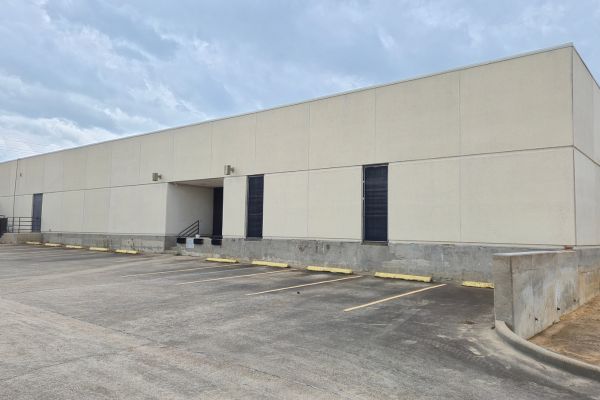 After Washing Services: The same warehouse building after receiving washing services. The dirt and stains have been removed from the walls, giving the surface a much cleaner and fresher appearance. The concrete base and parking lot remain unchanged, with the windows still covered with black grilles.