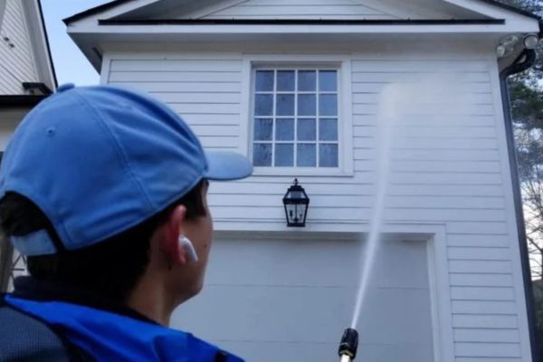 A person using a pressure washer to clean the exterior of a house, spraying water on the siding near a window, illustrating house washing services for exterior maintenance.