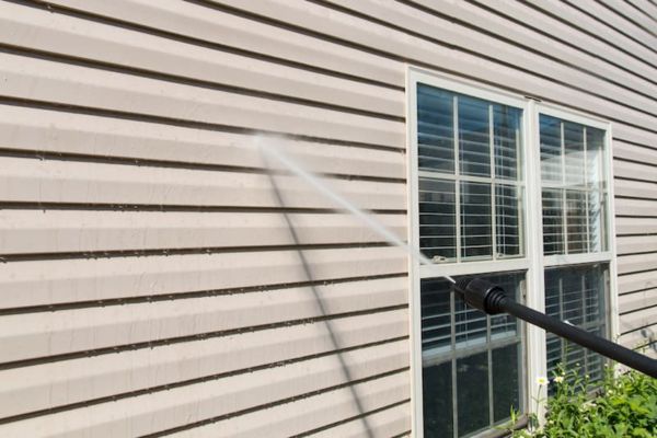 A pressure washer nozzle spraying water on the exterior siding of a house near a window, demonstrating house washing services to clean the building's surface.