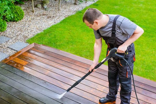 A person wearing protective gear using a pressure washer to clean a wooden deck in a backyard, demonstrating pressure washing services for outdoor wood surfaces.
