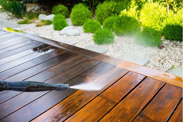 A pressure washer spraying water on a wooden deck, cleaning the surface as part of pressure washing services. The surrounding greenery and gravel path are visible in the background.