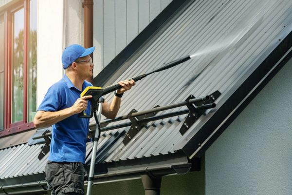 A person using a pressure washer to clean a metal roof, demonstrating roof washing services to remove dirt and grime from roofing materials.