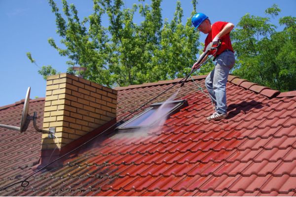 A person wearing safety gear using a pressure washer to clean the roof of a house, demonstrating roof washing services to remove dirt and debris from the roof tiles.