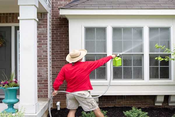 A person using a soft washing technique with a sprayer to clean the exterior of a house, focusing on the windows and walls as part of soft washing services.