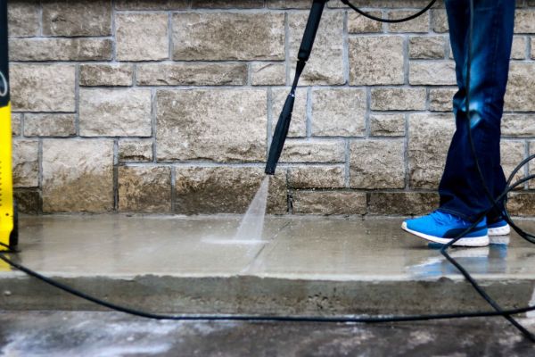 A person using a pressure washer to clean a concrete surface near a stone wall, demonstrating soft washing services to gently remove dirt and stains from outdoor surfaces.