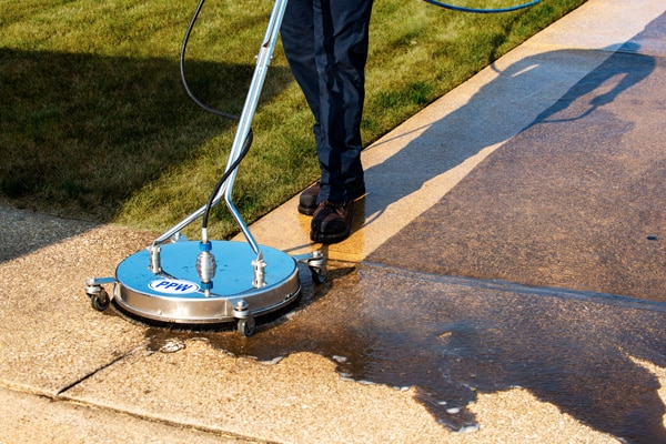 A person using a surface cleaner attachment on a pressure washer to clean a concrete sidewalk, demonstrating concrete washing services for a more efficient and even surface cleaning.