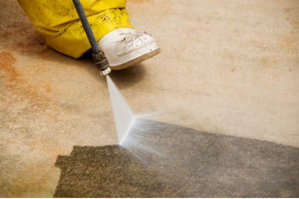 person wearing yellow protective gear using a pressure washer on a concrete surface, demonstrating concrete washing services to clean and restore the area.