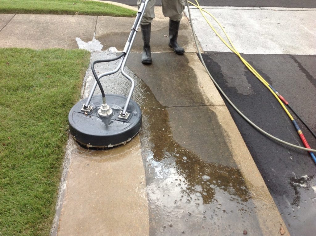 A technician is using a surface cleaner attachment on a pressure washer to clean a concrete sidewalk. The equipment helps to evenly wash the surface, removing dirt and stains. This is an example of concrete washing services designed to restore the cleanliness and appearance of paved areas.
