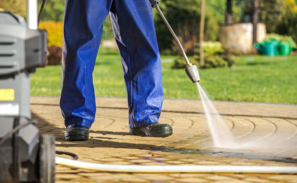 A worker in blue overalls using a pressure washer to clean a stone patio. The powerful water stream is effectively removing dirt and stains from the surface, highlighting the precision and efficiency of Professional Pressure Washing Services in restoring outdoor areas to their pristine condition.
