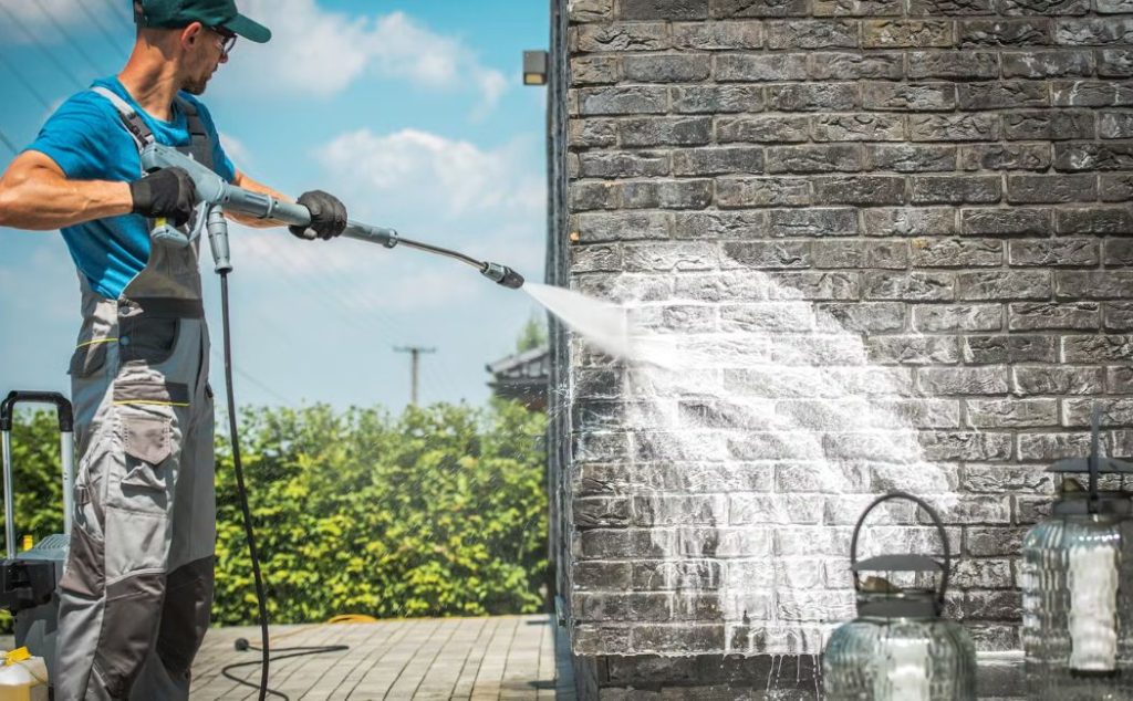 A professional wearing protective gear uses a high-pressure washer to clean a brick wall. The powerful water stream is removing dirt and stains, revealing the clean surface beneath. This image highlights the effectiveness and precision of Professional Pressure Washing Services for maintaining the exterior of buildings.