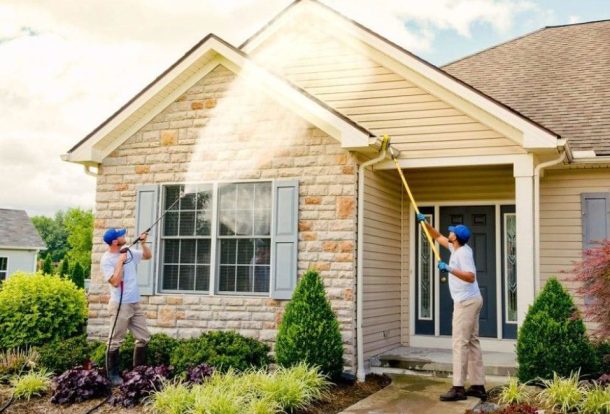 Two professionals are using pressure washers to clean the exterior of a home. One is using a long wand to reach the higher areas, while the other focuses on the lower section near the entrance. This scene highlights the effectiveness of pressure washing services.