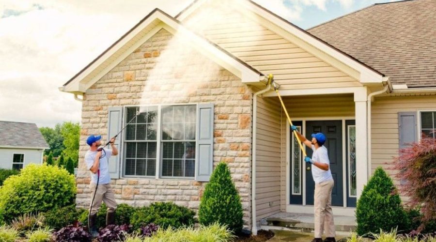 Two professionals are using pressure washers to clean the exterior of a home. One is using a long wand to reach the higher areas, while the other focuses on the lower section near the entrance. This scene highlights the effectiveness of pressure washing services.