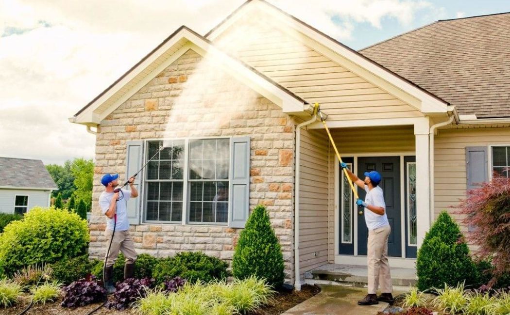 Two professionals are using pressure washers to clean the exterior of a home. One is using a long wand to reach the higher areas, while the other focuses on the lower section near the entrance. This scene highlights the effectiveness of pressure washing services.