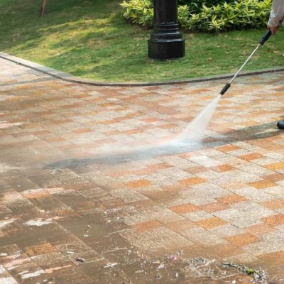 A professional wearing protective gear uses a pressure washer to clean a paved concrete surface. The powerful water stream is effectively removing dirt and debris, leaving the concrete surface looking fresh and clean. This image demonstrates the quality and effectiveness of Concrete Washing Services.