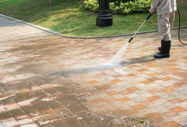 A professional wearing protective gear uses a pressure washer to clean a paved concrete surface. The powerful water stream is effectively removing dirt and debris, leaving the concrete surface looking fresh and clean. This image demonstrates the quality and effectiveness of Concrete Washing Services.