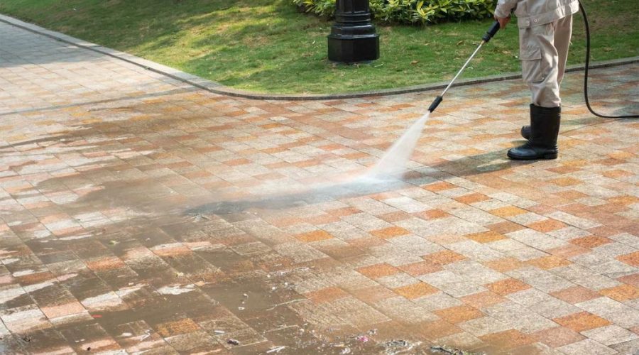 A professional wearing protective gear uses a pressure washer to clean a paved concrete surface. The powerful water stream is effectively removing dirt and debris, leaving the concrete surface looking fresh and clean. This image demonstrates the quality and effectiveness of Concrete Washing Services.