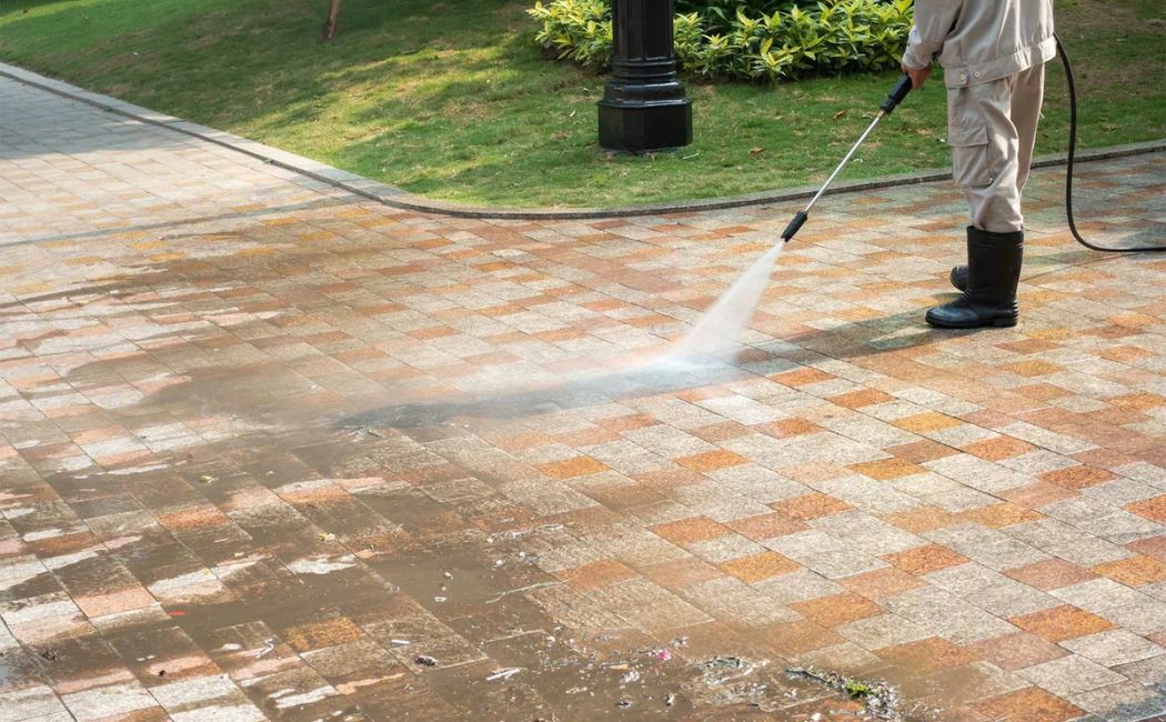 A professional wearing protective gear uses a pressure washer to clean a paved concrete surface. The powerful water stream is effectively removing dirt and debris, leaving the concrete surface looking fresh and clean. This image demonstrates the quality and effectiveness of Concrete Washing Services.