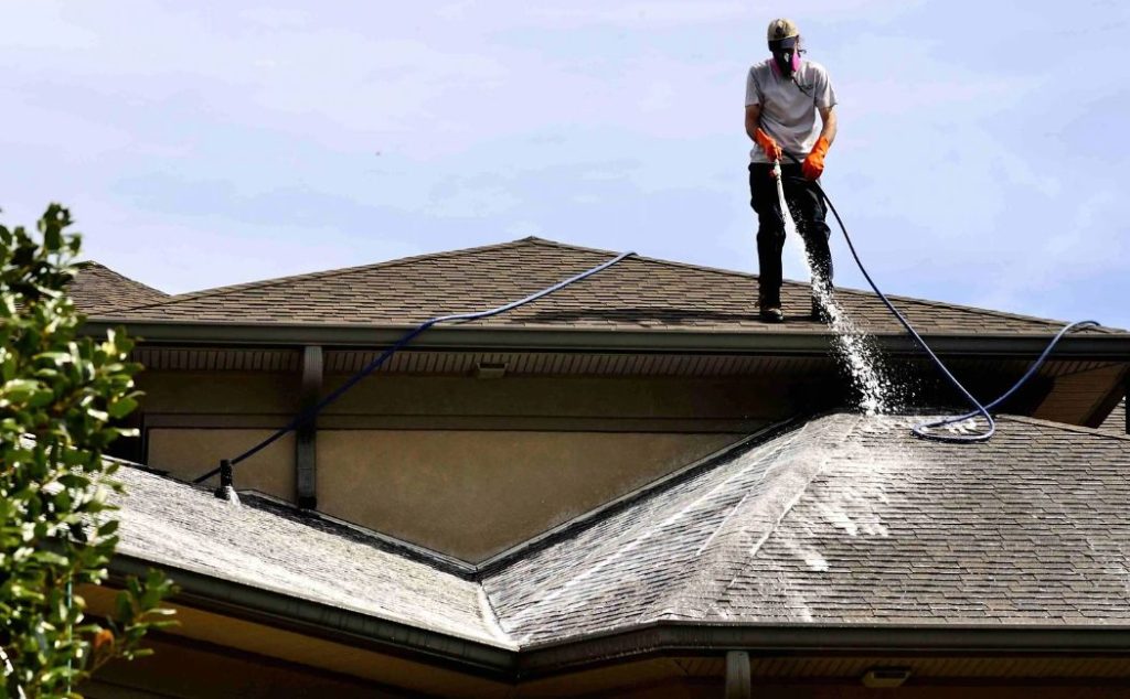 Roof washing services expert cleaning a shingle roof using safe water spraying techniques to remove dirt, algae, and debris.