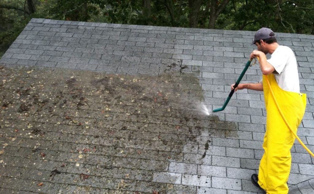 A person in yellow rain gear is using a pressure washer to clean moss and debris off a roof. The image highlights the effectiveness of professional roof cleaning services.