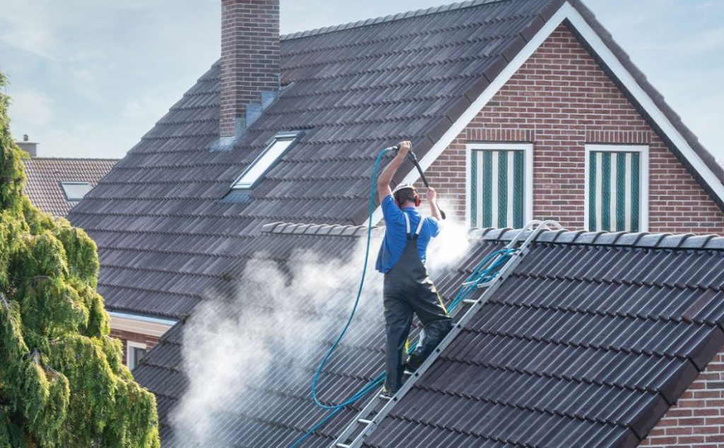 A professional is using a pressure washer to clean the roof of a house, with steam rising from the tiles. The worker is standing on a ladder and wearing safety gear to ensure a thorough and safe cleaning process.