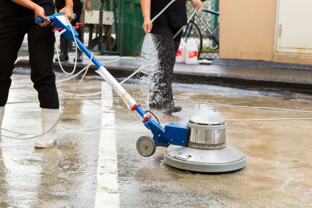 Professionals performing concrete washing services using industrial floor scrubbers and high-pressure water hoses to clean an outdoor concrete surface.