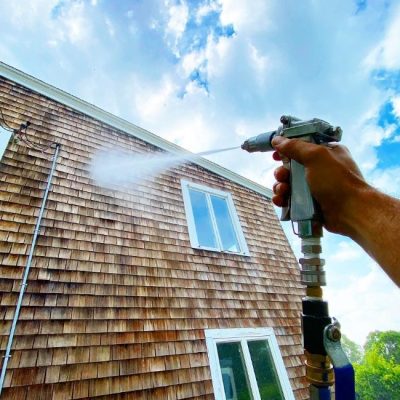 A person is using a pressure washer to clean the exterior of a house, focusing on the wood siding. The high-pressure water spray removes dirt and grime, helping to restore the home's appearance. This is an example of house washing services that enhance the curb appeal and cleanliness of residential properties.
