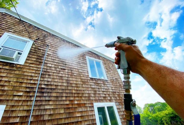 A person is using a pressure washer to clean the exterior of a house, focusing on the wood siding. The high-pressure water spray removes dirt and grime, helping to restore the home's appearance. This is an example of house washing services that enhance the curb appeal and cleanliness of residential properties.