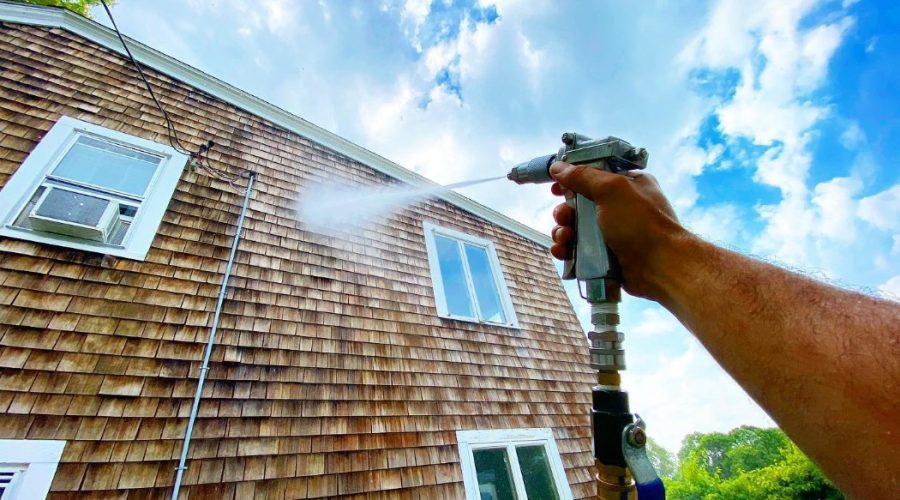 A person is using a pressure washer to clean the exterior of a house, focusing on the wood siding. The high-pressure water spray removes dirt and grime, helping to restore the home's appearance. This is an example of house washing services that enhance the curb appeal and cleanliness of residential properties.