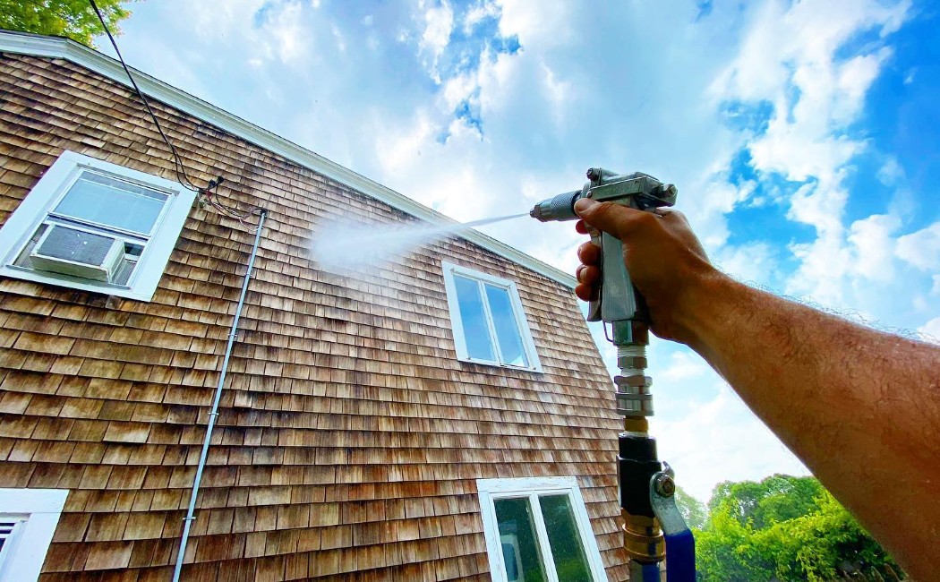 A person is using a pressure washer to clean the exterior of a house, focusing on the wood siding. The high-pressure water spray removes dirt and grime, helping to restore the home's appearance. This is an example of house washing services that enhance the curb appeal and cleanliness of residential properties.
