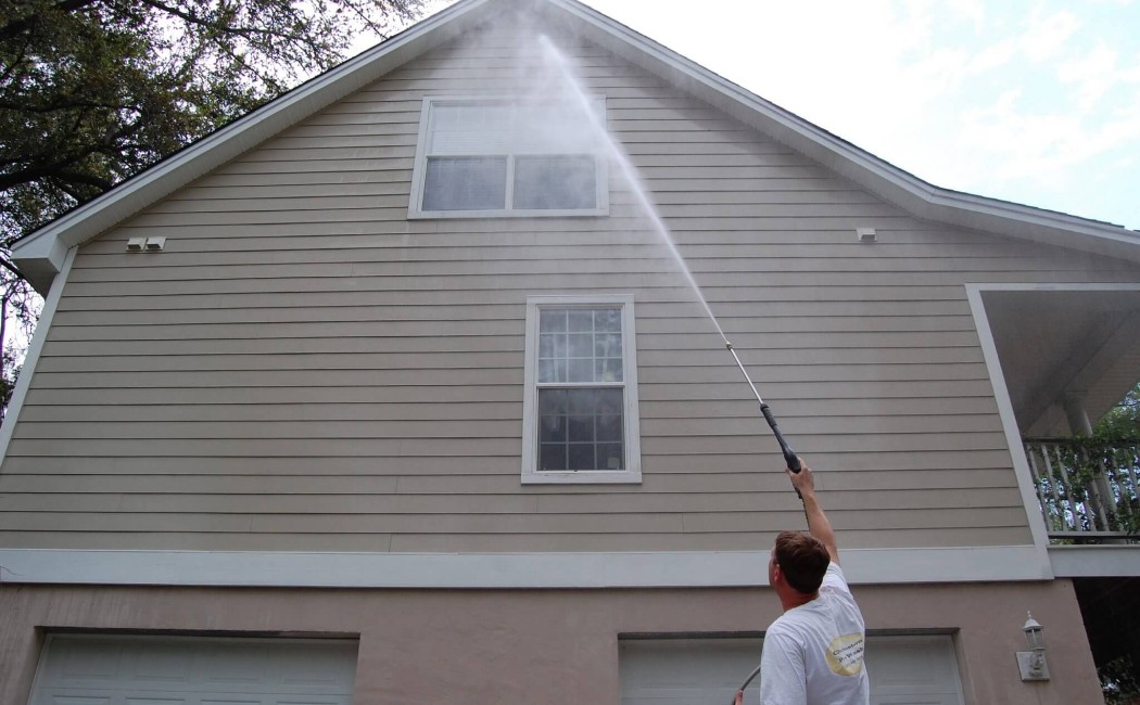 The image depicts a person using a pressure washer to clean the exterior of a house. Standing on the ground, the individual is aiming a powerful stream of water at the upper siding near the roofline, effectively removing dirt and grime. The clean, well-maintained surface demonstrates the results of professional house washing services.