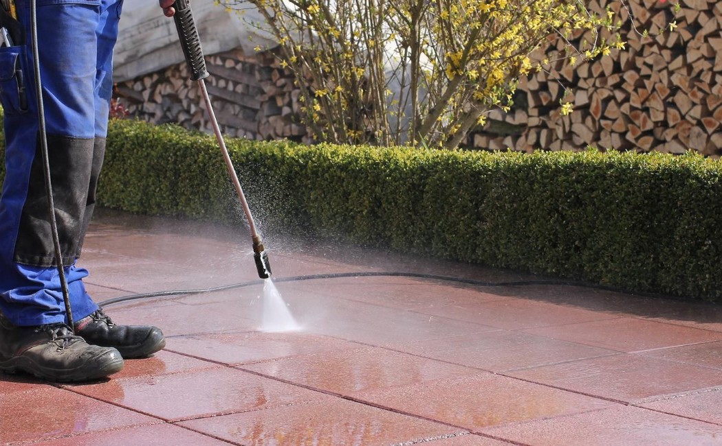 Worker using a soft washing system to clean red patio tiles in a neatly landscaped yard, showcasing the Best Soft Washing Services for outdoor surfaces.