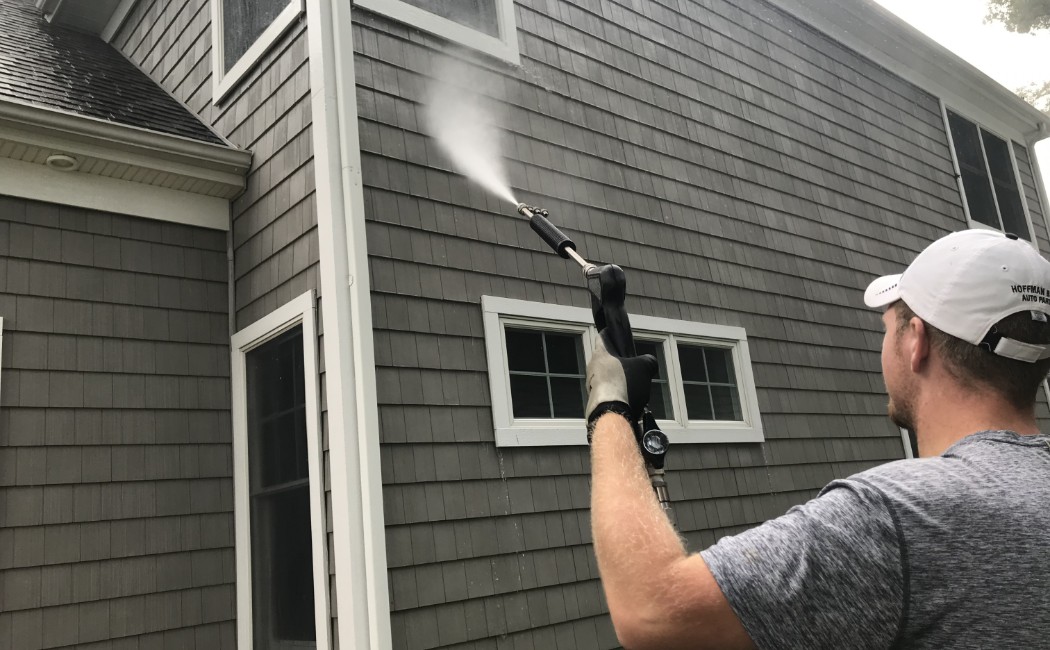 A person is using a pressure washer to clean the exterior of a house, targeting the siding with a strong water spray. The process helps to remove dirt, algae, and mildew, restoring the home's appearance.