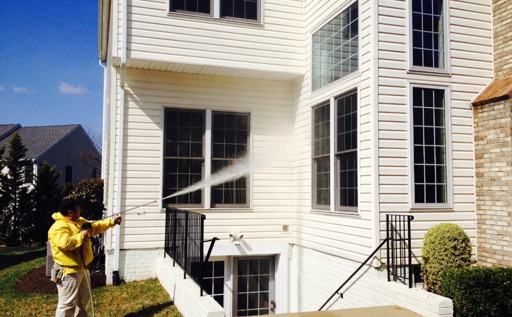 The image shows a professional cleaner using a pressure washer to clean the exterior of a house. Wearing a yellow jacket and beige pants, the technician is directing a powerful stream of water at the side of the house, targeting the windows and siding. The surrounding lawn and tidy garden suggest the area is well-maintained.