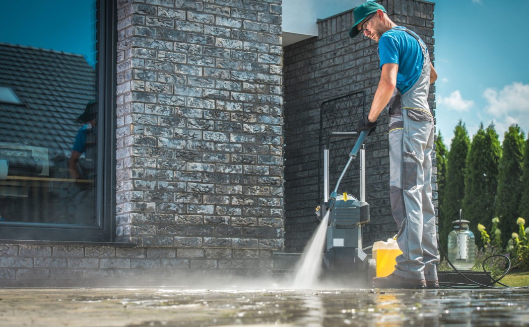 Professional technician performing exterior surface cleaning using a pressure washer on a modern home’s stone pavement under clear skies, representing the Best Soft Washing Services for residential properties.
