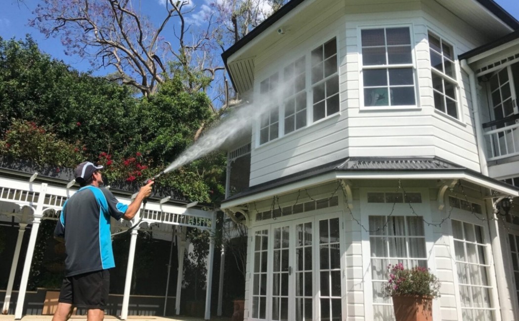 The image shows a professional house washing technician using a pressure washer to clean the upper part of a home's exterior. Wearing a cap and a blue and grey uniform, they aim the high-pressure water stream at the house's siding and windows, effectively removing dirt and grime. The surrounding lawn and well-maintained yard highlight the clean environment.