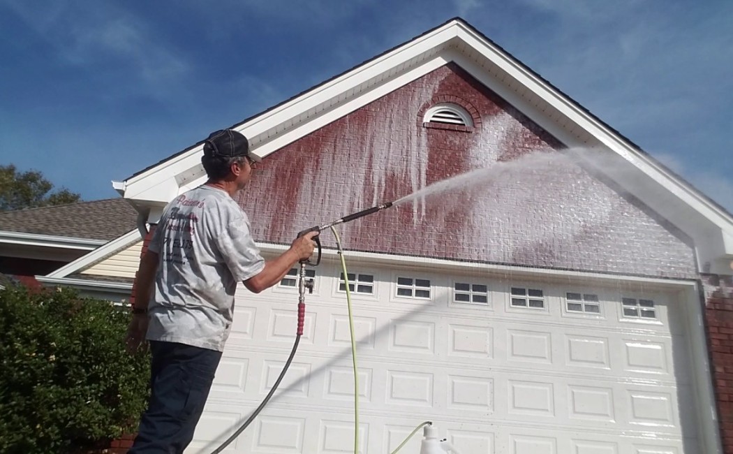 A professional from a roof washing company uses a pressure washer to clean the exterior of a home, removing dirt and grime from the brick surface.